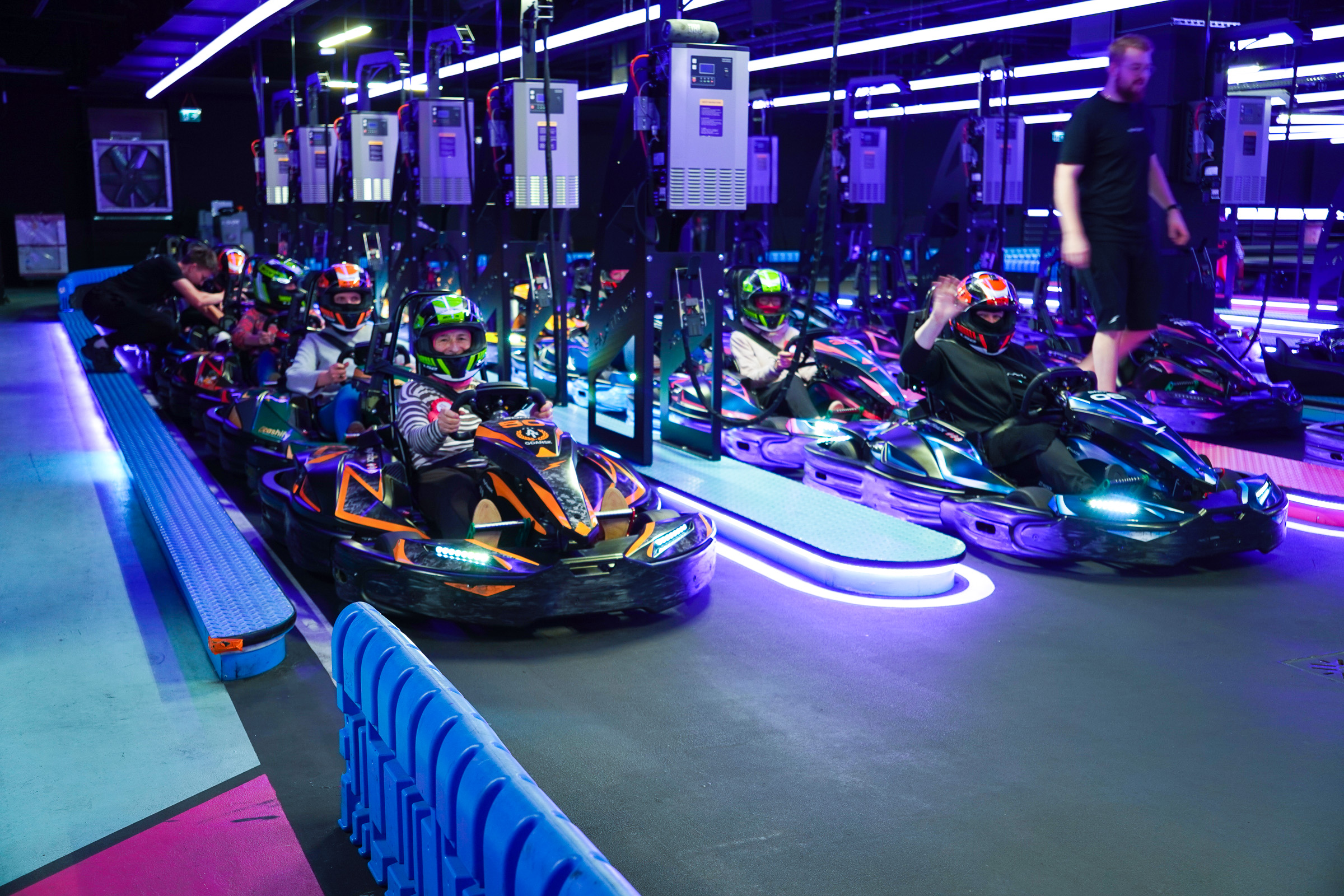 Uczestniczki projektu siedzą w gokartach ustawionych w rzędzie na torze Fast Track. W tle instruktor i neonowe oświetlenie.